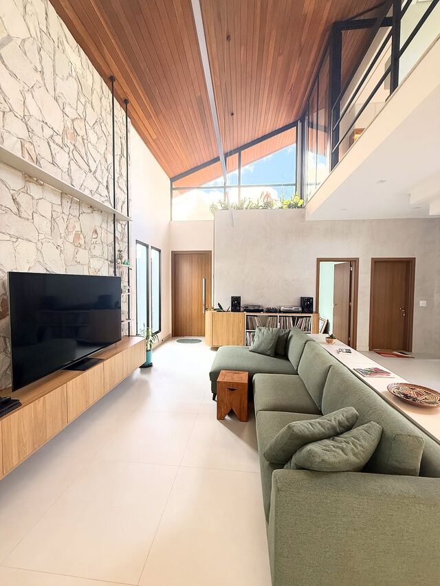 Sala ampla com pé-direito alto, sofá verde e parede de pedra